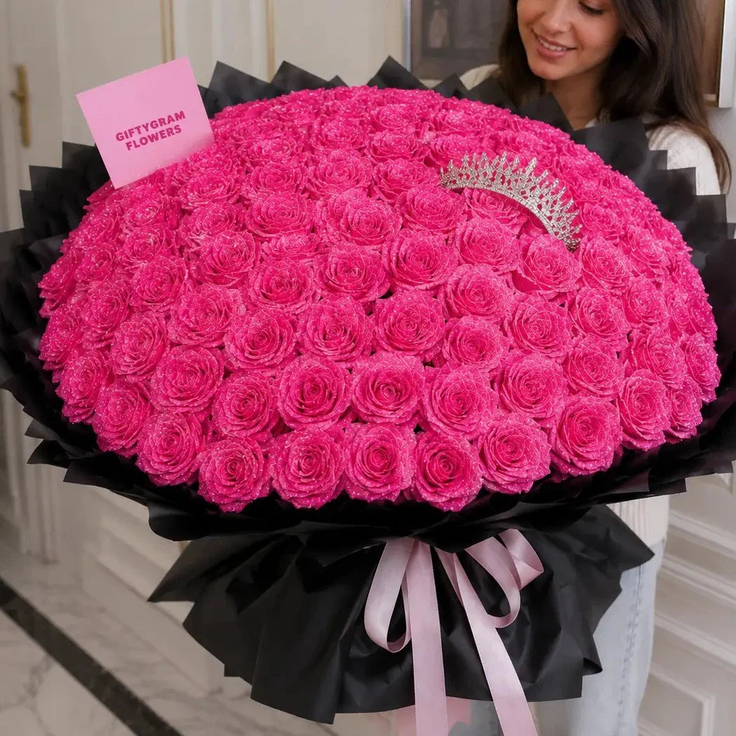 A large, round bouquet of bright pink roses wrapped in black paper with a light pink ribbon, held by a woman. A small pink card reading 'GIFTYGRAM FLOWERS' is attached, and a silver tiara rests on top.