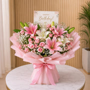 Birthday Flowers of pink lilies and white flowers with a 'Happy Birthday!' card on a marble surface.