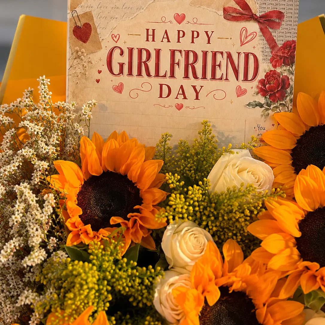 A floral arrangement for Happy Girlfriend Day featuring bright orange sunflowers, white roses, green filler flowers, and a printed card that reads “HAPPY GIRLFRIEND DAY” with red hearts and roses.