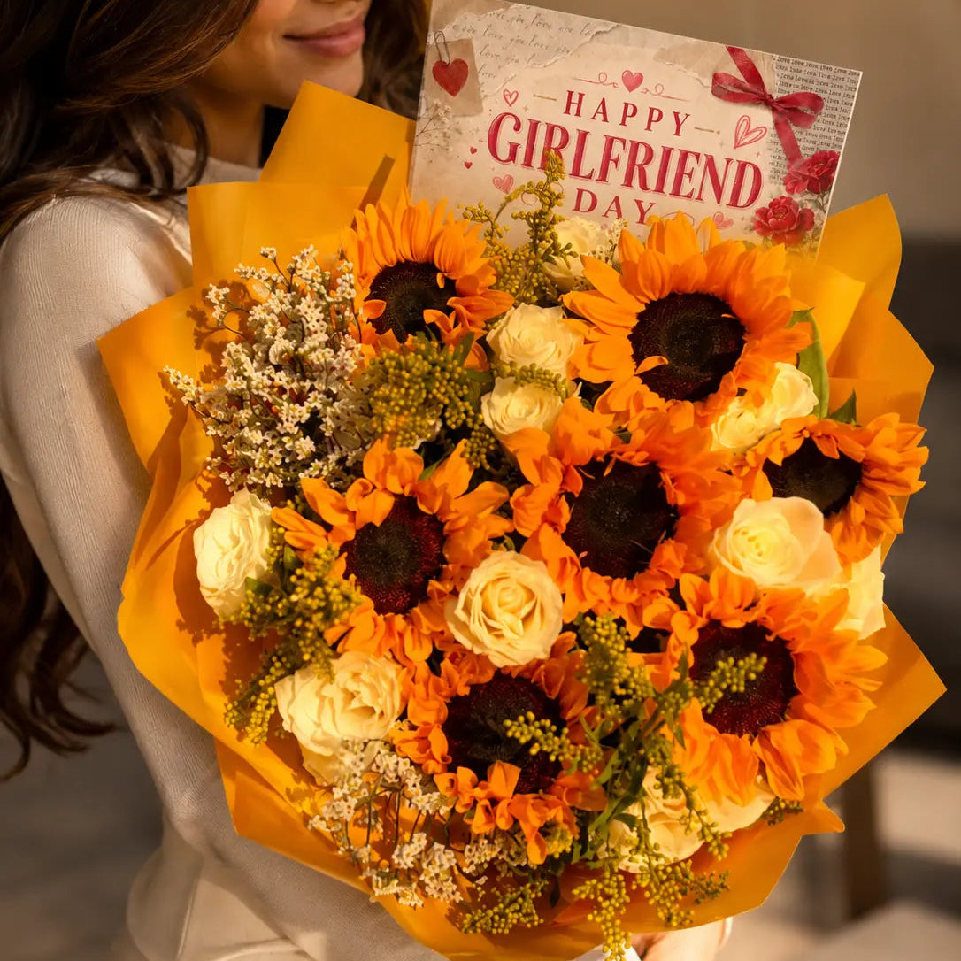 A woman in a white dress holds a large sunflower and rose bouquet wrapped in bright orange paper, with a card that reads 'Happy Girlfriend Day' visible at the top.