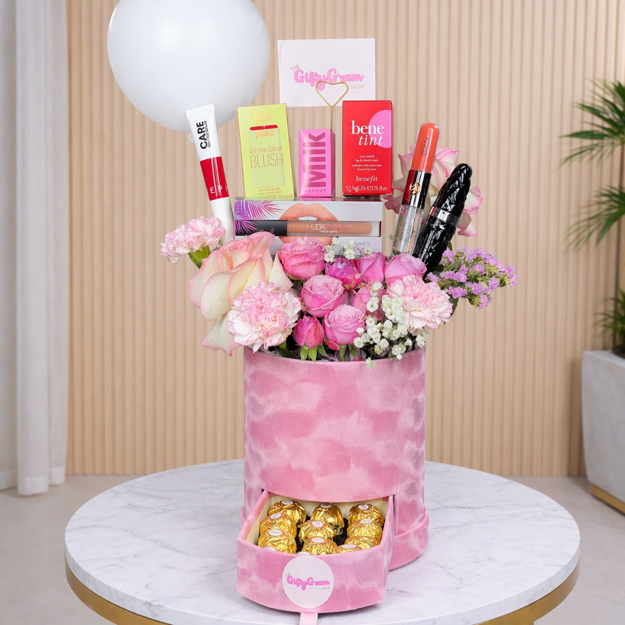 Pink floral arrangement with makeup items and chocolates on a marble surface.