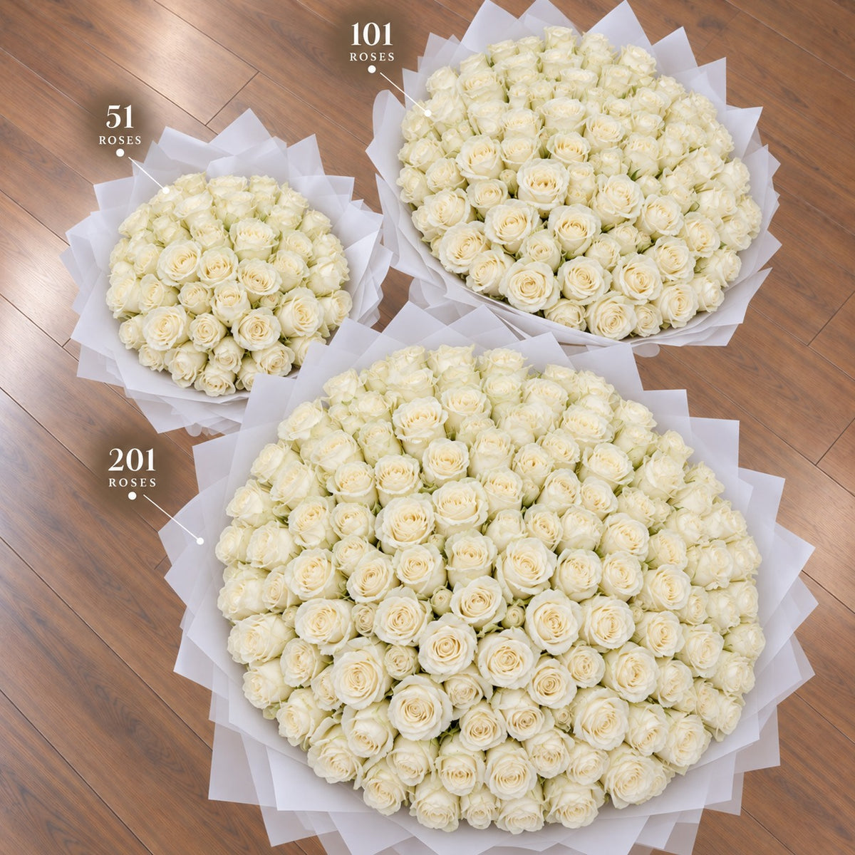 Three round arrangements of white roses on a wooden floor, labeled with counts 51, 101, and 201 roses. The large arrangement in the foreground is wrapped in white tissue paper.