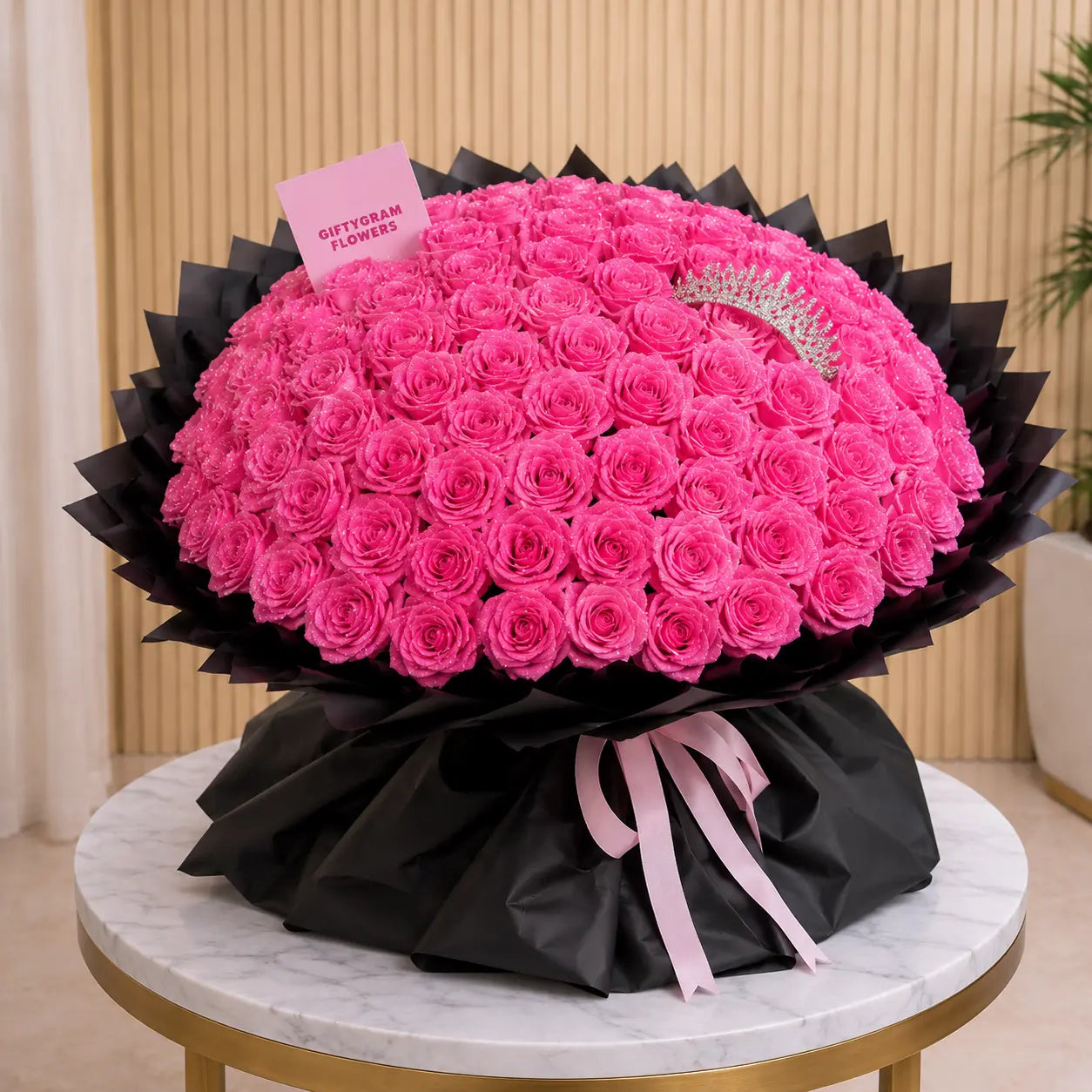 A large round bouquet of bright pink roses wrapped in black paper with a pink ribbon, displayed on a marble-topped table. A small card reading 'GiftyGram FLOWERS' is tucked into the bouquet, and a silver bow is tied at the back.