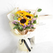 Bouquet of sunflowers and other flowers wrapped in brown paper with a black ribbon on a white background.