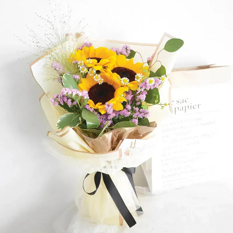 Bouquet of sunflowers and other flowers wrapped in brown paper with a black ribbon on a white background.