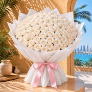 A large bouquet of white roses wrapped in white tissue paper with a pink ribbon bow, displayed on a wooden table with a beach and city skyline in the background.
